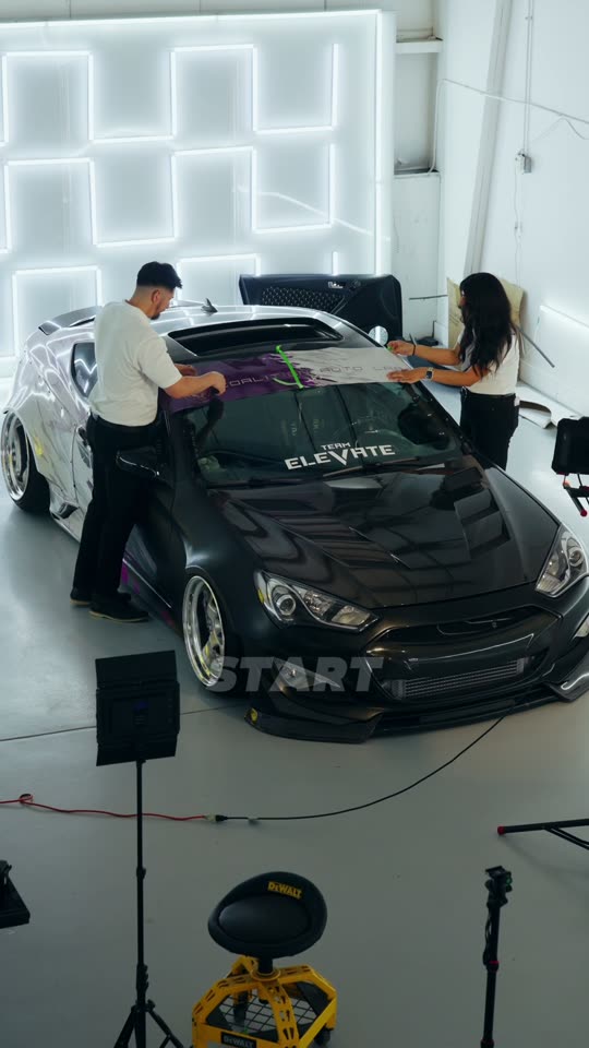 Two people fitting a shop banner to the windscreen of a black show car under studio lighting on a content day.