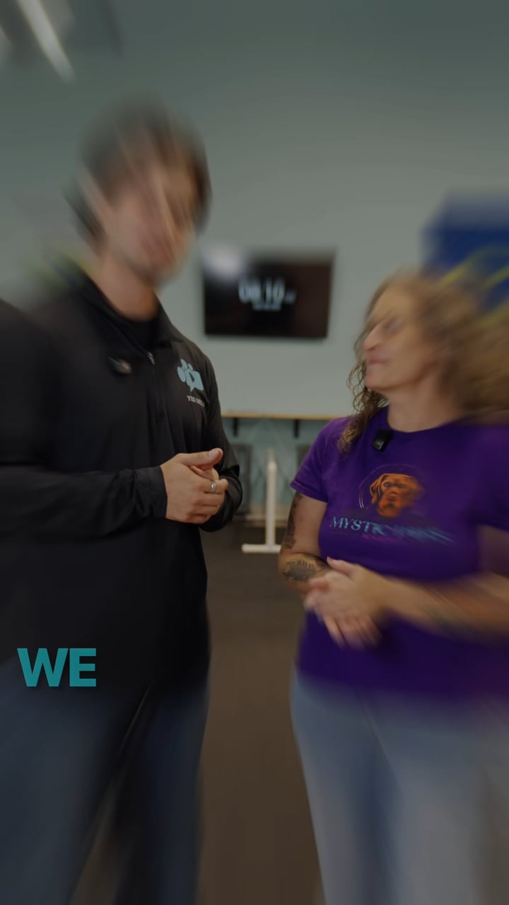 Reel still: a whip-pan frame of the owner of Mystic Paws Academy talking with Parker Yeo on the training floor, the word we in teal at the corner.